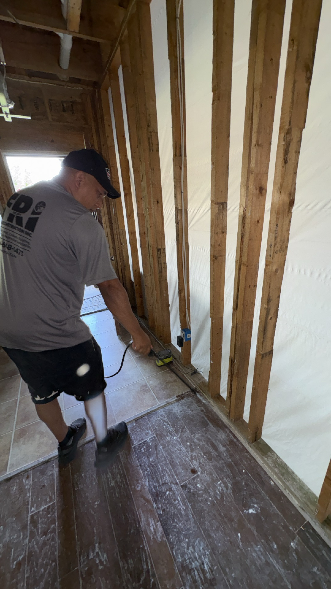 Worker cleaning and preparing water-damaged flooring in a hallway with exposed studs, using a specialized tool.
