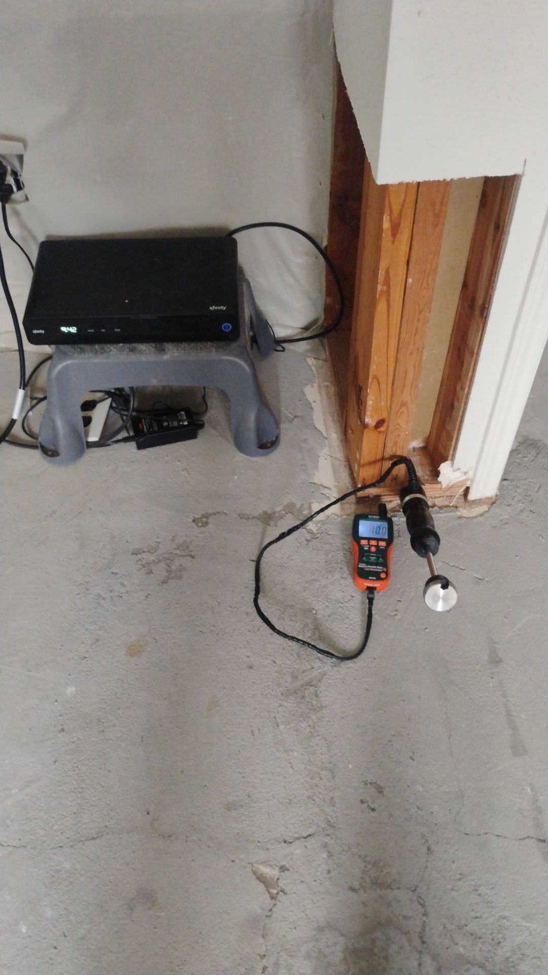 Moisture meter with pin probes testing moisture levels in a concrete floor next to an exposed wall stud during water damage assessment.